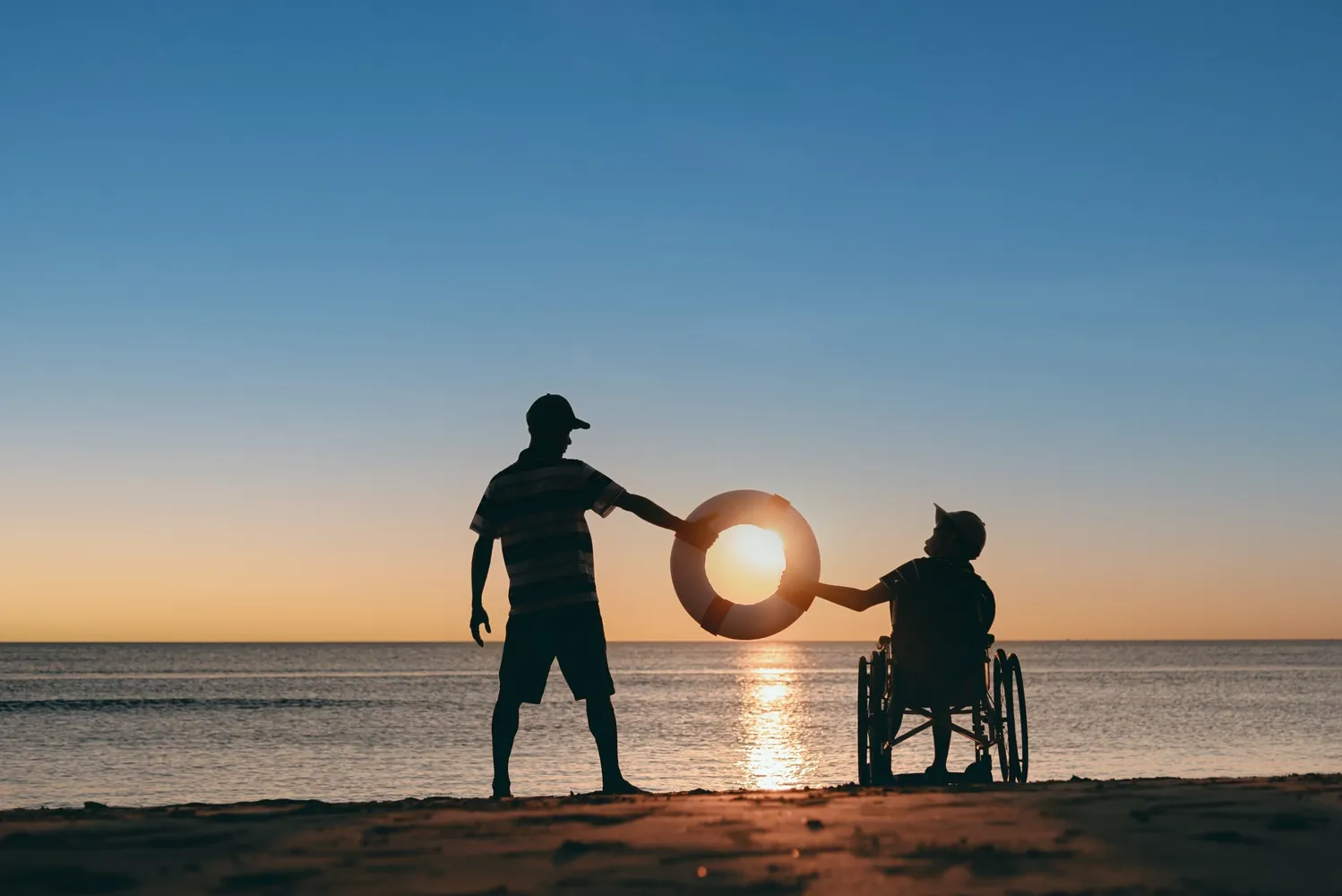 Two people on a beach one sitting in a wheelchair and the other standing, they are looking toward each other as they hold a round marine life preserver ring between them with the sun setting over the calm water glowing just off center of the life preserver ring.